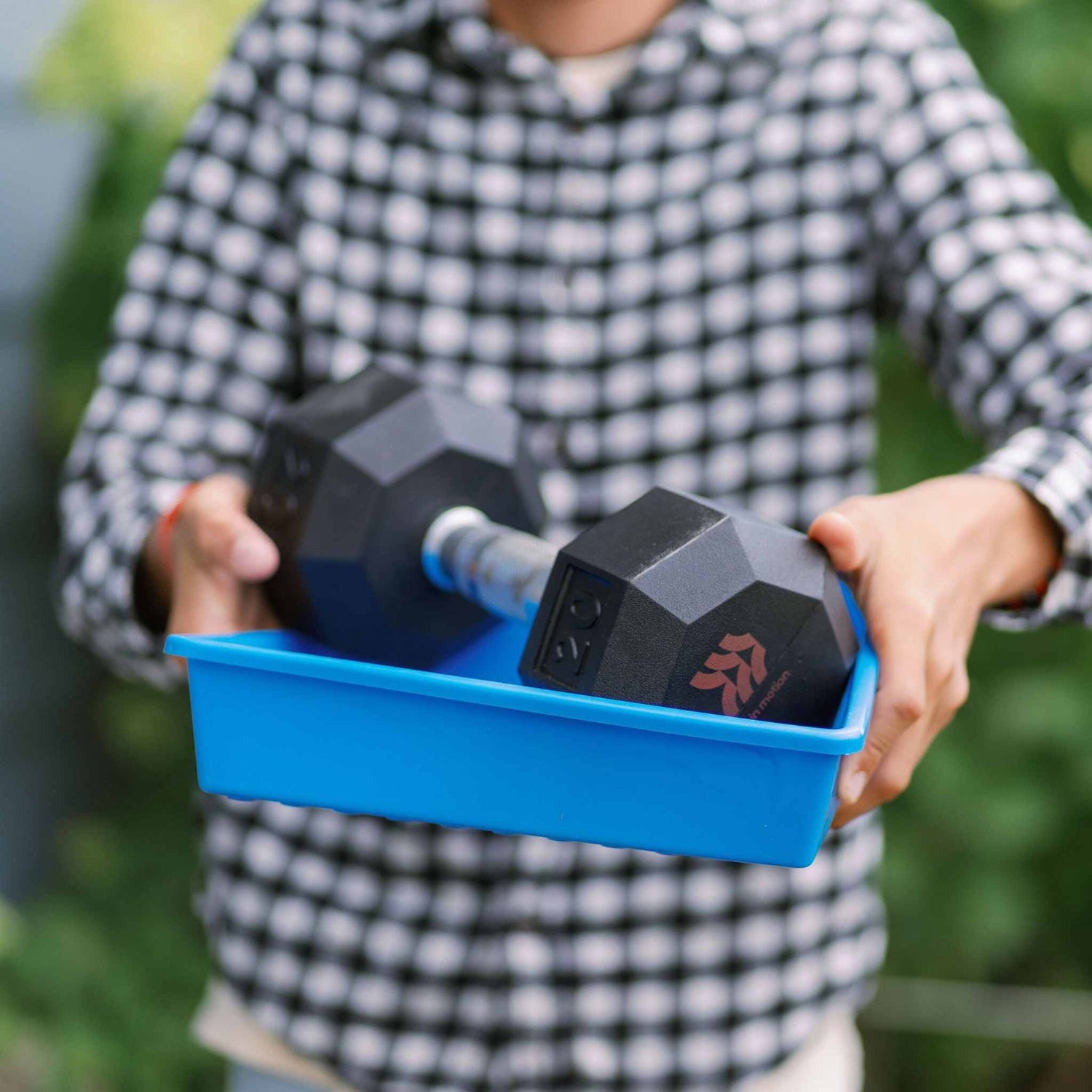 Person holding a blue container with dumbbells, wearing a checkered shirt outdoors. 