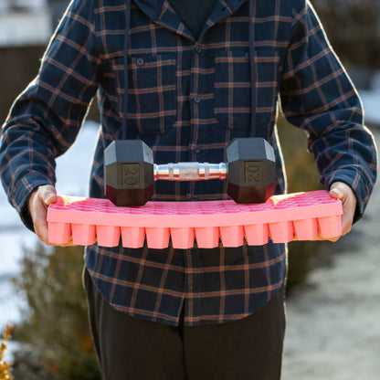 Person holding a pink propagation tray with a 20 lb. dumbbell on top, outdoors. 